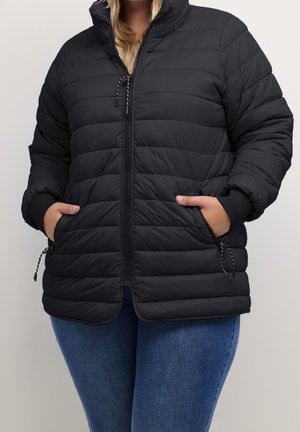 Osoba ubrana w czarną pikowaną kurtkę zapinaną na zamek błyskawiczny z rękami w kieszeniach zamykanych na zamek, w połączeniu z niebieskimi dżinsami na jednolitym tle.