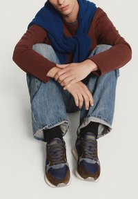 Zapatillas deportivas en tonos azul marino y marrón, con texturas de ante y malla, que presentan un collar acolchado y suela de goma, combinadas con jeans cortados y un suéter azul.