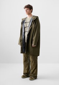 Olivengrüne wasserdichte Jacke mit Kapuze, schwarzer Innenfutter und Zugverschluss. Kombiniert mit passenden olivfarbenen Hosen und beigem Schuhwerk.