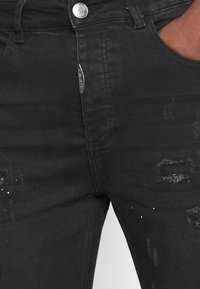 Primer plano de unos jeans negros desgastados con botón y cremallera plateados, llevados por una persona con la mano descansando en el bolsillo.