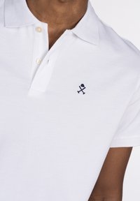 Camisa polo blanca con tejido texturizado, que presenta una tapeta de dos botones y un pequeño logotipo bordado en azul marino de artículos cruzados en el pecho.