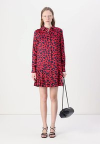 Abito button-up rosso e blu navy con motivo a cuori, gonna plissettata, maniche lunghe e colletto. Il modello tiene in mano una borsa nera.