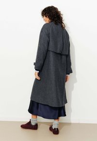 Manteau long à carreaux gris foncé avec un dos en couches, associé à une robe en satin bleu et des chaussures bordeaux. Comprend des manches roulées et un détail de bouton.