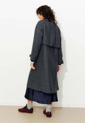Manteau long à carreaux gris foncé avec un dos en couches, associé à une robe en satin bleu et des chaussures bordeaux. Comprend des manches roulées et un détail de bouton.