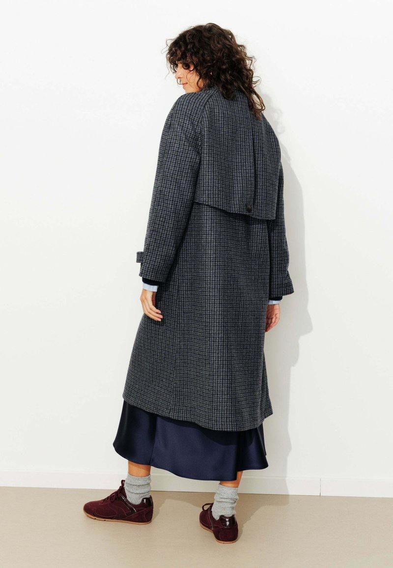 Manteau long à carreaux gris foncé avec un dos en couches, associé à une robe en satin bleu et des chaussures bordeaux. Comprend des manches roulées et un détail de bouton.