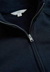 Jūras spēku zip-up džemperis, izgatavots no mīksta auduma, ar sudraba rāvējslēdzēju un etiķeti ar uzsaitu "THE SET" un izmēru "M" apkaklē.