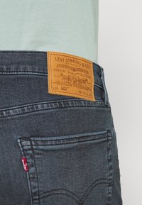 Mörkblå denimjeans med brun läderlapp med logotyp och detaljer. Inkluderar bakficka med röd etikett som visar sömdesign.