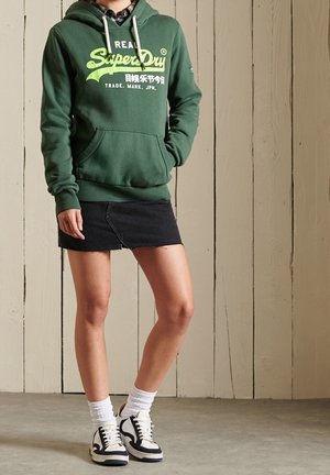 Sudadera con capucha verde con logo frontal, usada sobre una falda de mezclilla negra. Combinada con calcetas blancas y zapatillas negras y blancas. Fondo de paneles de madera.