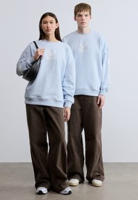 Ανοιχτό μπλε φούτερ με κεντημένο λογότυπο, συνδυασμένο με καφέ παντελόνια με φαρδιά μπατζάκια. Ένα μοντέλο κρατάει μια μαύρη τσάντα, και οι δύο φοράνε λευκά αθλητικά παπούτσια.