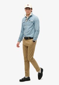 Lichtblauw denim overhemd met knoopsluiting, khaki chino's, zwarte riem, zwarte instappers en een crème pet met een logo. Taille- en casual pasvorm.