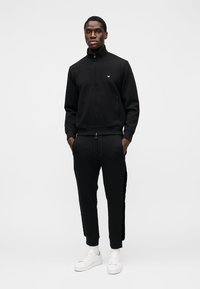 Svart treningsdress med glidelås og lommer, med høy krage og tettsittende mansjetter, kombinert med hvite joggesko for et komplett atletisk utseende.