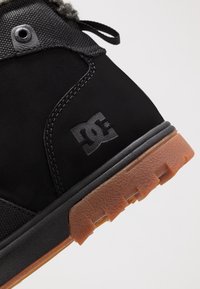 Czarny but z cholewką z teksturowanej tkaniny i gumową podeszwą. Posiada logo DC, wzmocnione szwy oraz wzorzysty gumowy spód w kolorze jasnobrązowym.