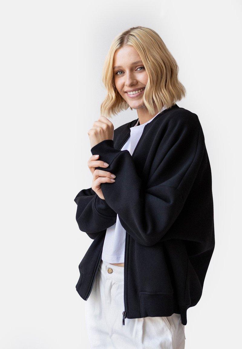 Czarna bomberka zapinana na zamek z prążkowanymi mankietami, luźny krój i miękka tekstura. Połączona z białą bluzką i beżowymi spodniami.