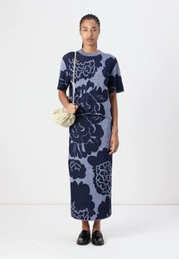 Costum din două piese din tricot în nuanțe de albastru, cu un model floral. Bluză cu mânici scurte și fustă lungă, asortată cu o geantă bej deschis și pantofi negri.
