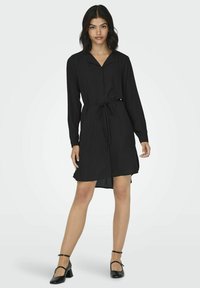 Abito camicia nero a maniche lunghe con colletto, cinturino da annodare, taglio dritto e spacco laterale, realizzato in un tessuto leggero e liscia.