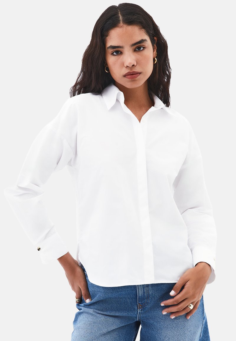 Camisa blanca de botones con cuello, mangas largas y detalles en los puños. Llevada con jeans de color azul claro, de corte relajado.