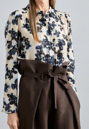 Mujer con una blusa crema con patrones florales en negro y azul, combinada con pantalones marrones de cintura alta atados con un cinturón de tela.