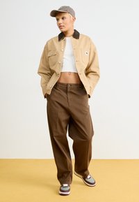Kaki utilityjacka med mörk krage, vit croptopp, bruna lösa byxor och flerfärgade sneakers. Modellen poserar mot en neutral bakgrund.