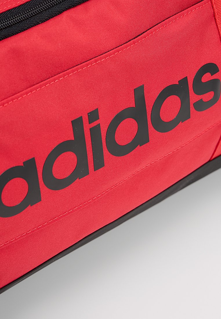 Mochila de tela roja con una cremallera negra y un prominente logo negro de "adidas" en la parte frontal. Presenta una textura suave y un diseño plano.