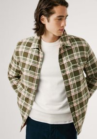 Camisa de franela a cuadros verdes con acentos en blanco y rojo, con botones en la parte delantera, dos bolsillos en el pecho, llevada sobre un suéter blanco y pantalones oscuros.
