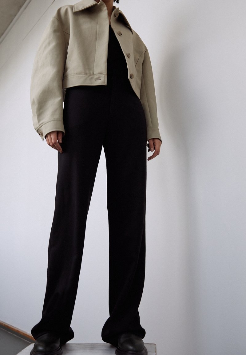 Personne portant une veste courte beige, un pantalon noir à jambes larges et des chaussures noires, debout devant un mur blanc uni.