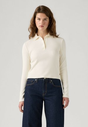 Ung kvinde med bølget brunt hår iført en cremefarvet poloshirt med lange ærmer og mørkeblå højtaljede jeans foran en ensfarvet baggrund.