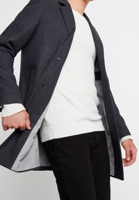 Uomo che indossa un cappotto grigio scuro sopra un maglione bianco testurizzato e pantaloni neri, tenendo il cappotto aperto per mostrare la fodera interna.