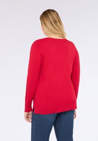 Žena s blond vlasy, oblečená v červeném svetru s dlouhým rukávem a modrých džínech, stojí zády k nám na jednoduchém světlem pozadí.