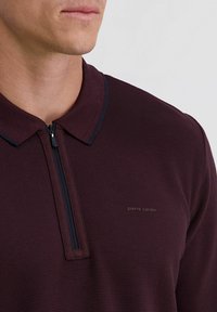 Gros plan sur un homme portant un polo zippé bordeaux foncé avec col et un petit logo "pierre cardin" sur la poitrine.