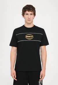 Černé tričko s krátkým rukávem z bavlny, s zlatým logem EA7 na hrudi a bílými vodorovnými pruhy přes přední část.