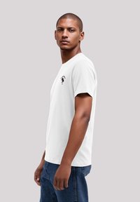 Weißes Baumwoll-T-Shirt mit kurzen Ärmeln, das auf der oberen linken Seite ein schwarzes Grafikdesign eines Palmenbaums zeigt. Trägt dazu eine blaue Jeans.