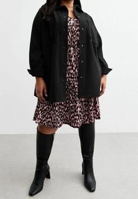 Schwarze oversize Jacke mit großen Knöpfen, getragen über einem rosa Leopardenmuster-Kleid. Knielange schwarze Stiefel runden das Outfit ab.