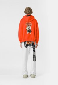 Sudadera roja que presenta un gráfico de un oso de dibujos animados con un traje de karate. Combinada con pantalones blancos que muestran texto y patrones en negro. Texturas suaves visibles.