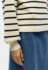 Maglione crema lavorato a maglia con righe orizzontali nere, con polsini a coste, abbinato a una gonna in denim blu. Mano visibile che poggia sulla gonna.