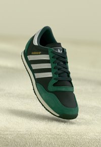 Green and black Adidas sneaker with three silver stripes, textured suede and mesh upper, rubber sole, and gold logo accent.