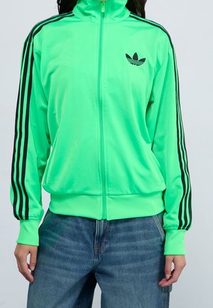 Osoba nosząca jaskrawozieloną bluzę sportową Adidas zapinaną na zamek, z czarnymi paskami na rękawach i logo na piersi, w połączeniu z niebieskimi dżinsami.