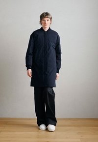 Námornícky modrý vatovaný kabát s vysokým golierom, zapínaný na predný zips a s elastickými manžetami, nosený cez voľné čierne nohavice a biele tenisky. Svetlá drevená podlaha.