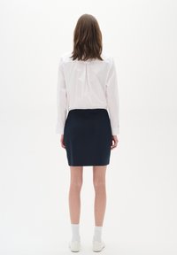 Biała koszula z długim rękawem i plisą na plecach, zestawiona z granatową spódnicą ołówkową. Modelka nosi białe skarpetki i minimalistyczne białe buty.