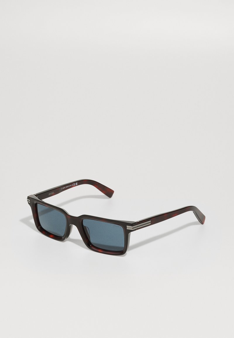 Sunglasses with a glossy dark tortoiseshell frame, square lenses, blue tint, and metal accents on the brow. Textured temples provide grip.