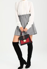 Žena v bílé blůze, šedé mini sukni a černých botách nad kolena, držící černou a červenou kabelku se zlatou sponou před bílým pozadím.