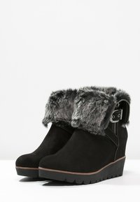 Černé surové klínové boty s šedou kožešinovou manžetou, bočním zipem a texturovanou gumovou podrážkou. Obsahují ozdobný pásek kolem kotníku a kovové akcenty.