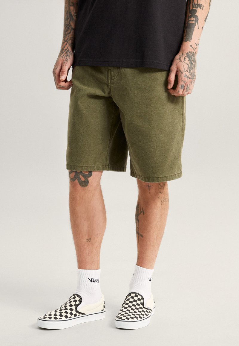 Shorts vert olive avec une coupe classique, en coton, et avec des ourlets roulés. Associés à des chaussures de type slip-on à carreaux noirs et blancs et des chaussettes blanches.
