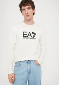 Langermet kremfarget T-skjorte med svart "EA7 EMPORIO ARMANI" logo, kombinert med lyseblå jeans. Mykt bomullsmateriale, avslappet passform.