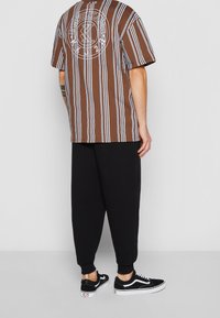 Muž v černých kalhotách, černo-bílých teniskách a hnědém tričku s krátkým rukávem, které má bílé svislé pruhy a bílý kulatý design na zádech.