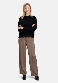 Czarny dzianinowy sweter z koronkową lamówką, zestawiony z jasnobrązowymi szerokimi spodniami i czarnymi botkami za kostkę. Modelka stoi ze skrzyżowanymi ramionami.