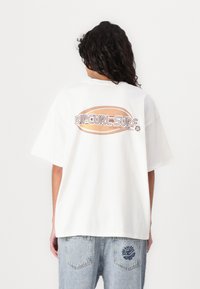 Camisa de t-shirt de gran tamaño en blanco con un gran gráfico en texto naranja y marrón que dice "RIPCURL-SURF" en la parte posterior. Tejido de algodón suave.