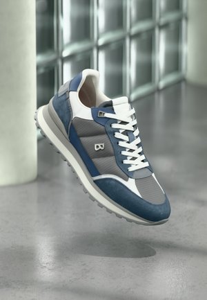 Stilīgas zilas, pelēkas un baltas sporta kurpes ar baltām auklām, kas lidinās virs pulēta betona grīdas stikla bloku sienas fonā.