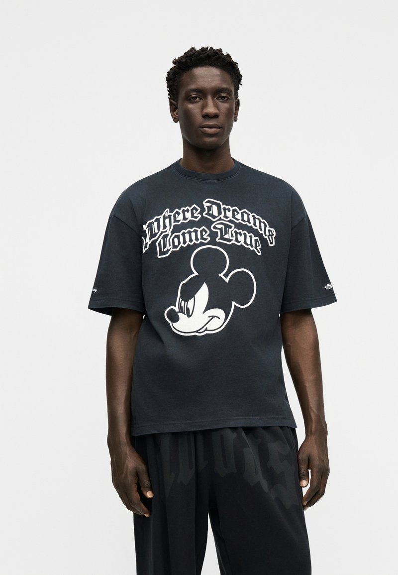 Mand iført sort oversized T-shirt med hvid Mickey Mouse-grafik og teksten "Where Dreams Come True" på fronten, der står foran en ensfarvet baggrund.