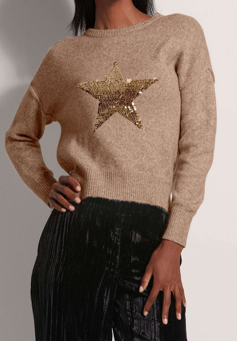 Beige neulepusero, jossa etuosassa kultainen paljettitähtikuosi. Resorireunus helmassa ja hihansuissa, yhdistetty mustiin, teksturoiduihin housuihin.