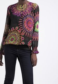 Blouse noire en voile transparent avec des motifs floraux multicolores vibrants, manches longues avec poignets à volants et encolure ronde avec boutons.